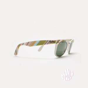 May include: White-framed sunglasses with green lenses. The temples feature a striped pattern in shades of pink, green, and tan. The Ray-Ban logo is visible on the temple and lens.