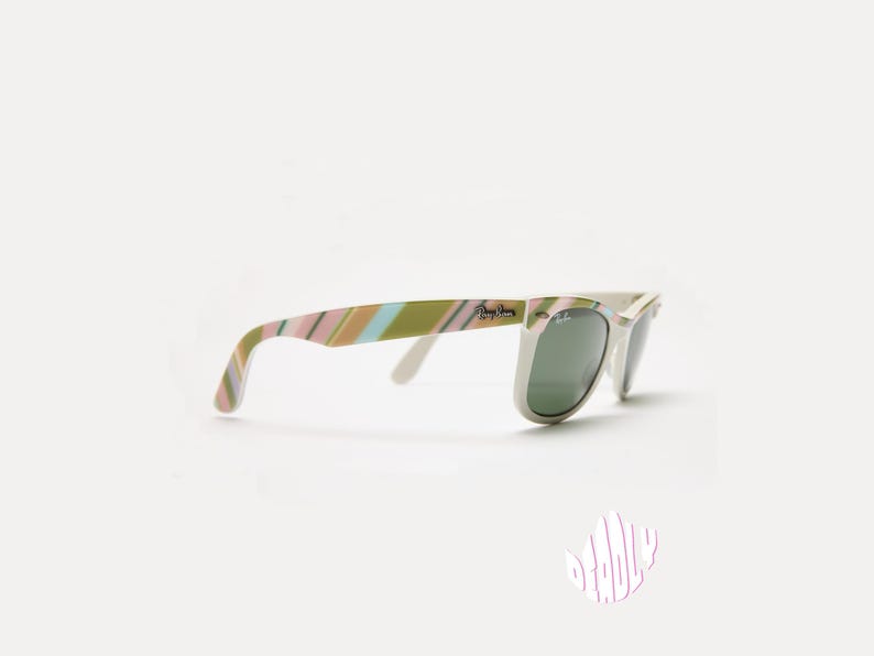 May include: White-framed sunglasses with green lenses. The temples feature a striped pattern in shades of pink, green, and tan. The Ray-Ban logo is visible on the temple and lens.