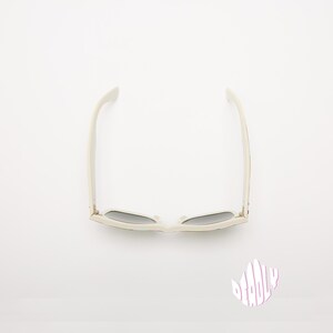 May include: Off-white sunglasses with dark lenses, viewed from above. The sunglasses have a classic rectangular frame and are set against a white background. The image also includes a small, stylized graphic with the word "DEADLY".