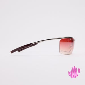 May include: A pair of rimless sunglasses featuring a silver metal frame and gradient red lenses that transition from a darker shade at the top to a lighter shade at the bottom. The temples are thin with dark brown tips. The word DEADLY is visible.