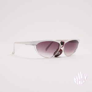 May include: Cat-eye sunglasses featuring a translucent clear frame and gradient lenses that transition from a darker tint at the top to a lighter shade at the bottom. A small dark tag is attached to the bridge. The word DEADLY is printed in pink.