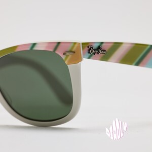 May include: Close-up of Ray-Ban sunglasses with a white frame and green lenses. The temples feature a colorful striped pattern in pink, green, and blue. The Ray-Ban logo is visible on the temple.