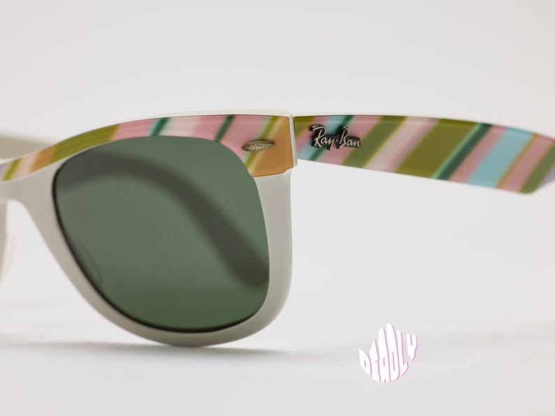 May include: Close-up of Ray-Ban sunglasses with a white frame and green lenses. The temples feature a colorful striped pattern in pink, green, and blue. The Ray-Ban logo is visible on the temple.