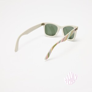 May include: White sunglasses with green lenses. The arms feature a colorful striped pattern in shades of pink, green, and yellow. The sunglasses are set against a white background, showcasing their design.