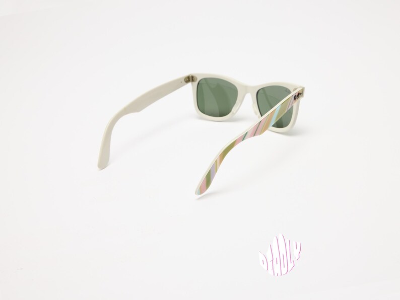 May include: White sunglasses with green lenses. The arms feature a colorful striped pattern in shades of pink, green, and yellow. The sunglasses are set against a white background, showcasing their design.