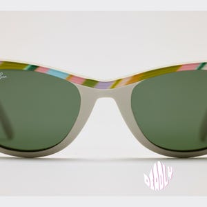 May include: Gray sunglasses with green lenses and a colorful striped pattern on the top frame. The Ray-Ban logo is visible on the upper left lens. The sunglasses have a classic, retro style.