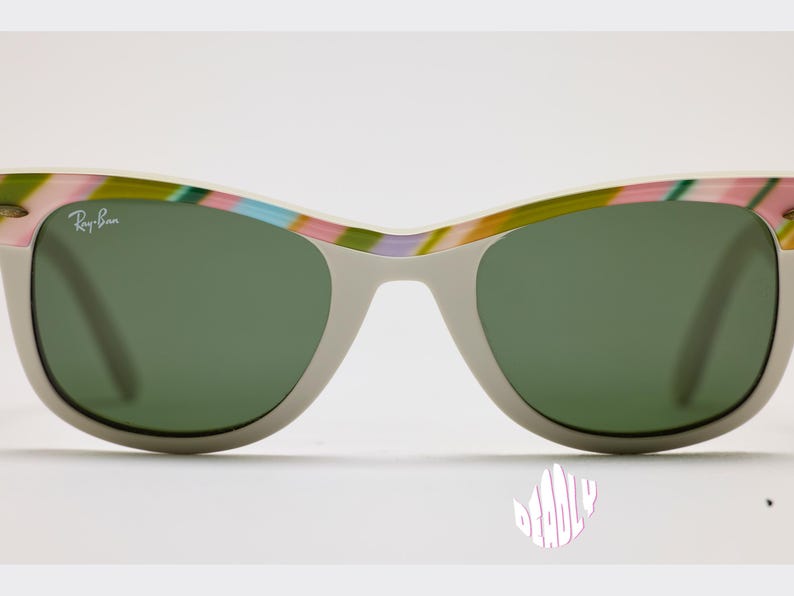 May include: Gray sunglasses with green lenses and a colorful striped pattern on the top frame. The Ray-Ban logo is visible on the upper left lens. The sunglasses have a classic, retro style.