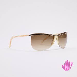 Könnte beinhalten: Randlose Sonnenbrille mit einem goldfarbenen Metallrahmen, einem dünnen Steg über der Nase und braunen Verlaufsgläsern, die nach unten hin heller werden. Die Bügel sind schmal und goldfarben, und das Wort DEADLY ist in Pink abgebildet.
