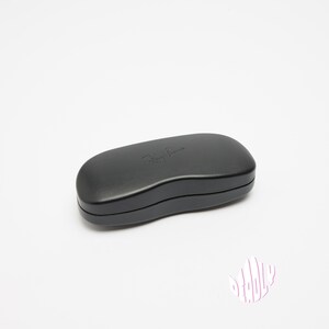 May include: A black, oval-shaped sunglasses case with the Ray-Ban logo embossed on the top. The case is closed and sits on a white surface. The case is approximately 6 inches long.