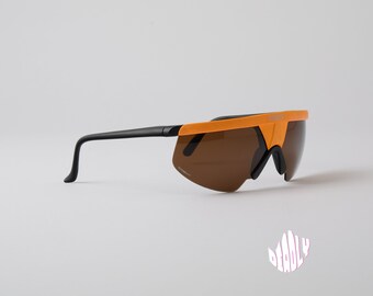 Puede incluir: Gafas de sol envolventes de color naranja y negro con lentes marrones. Las gafas tienen un diseño futurista.