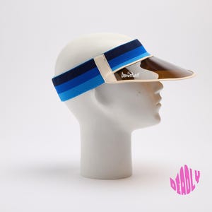 Peut inclure: Une visière de soleil dotée d'une large bande élastique à rayures horizontales dans des tons bleu foncé, bleu moyen et bleu clair. La visière possède un bord en plastique teinté marron translucide avec une bordure crème et le texte baruffaldi.