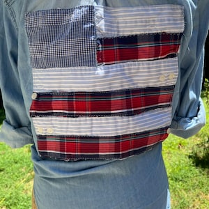 May include: A light blue denim shirt featuring a rectangular patch on the back designed to resemble an American flag. The patch is constructed from horizontal strips of blue gingham, red and blue plaid, and light blue and white striped fabric.