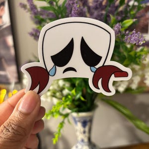 May include: A white sticker with a cartoon crab design. The crab has a sad face with black eyes and tears, and maroon claws. The sticker is held in front of a vase of flowers.