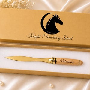 Puede incluir: Un abrecartas de metal dorado con mango de madera clara grabado con el nombre Valentina, presentado en una caja de regalo rectangular de color canela. La tapa de la caja muestra un gráfico negro de una cabeza de caballo en una luna creciente y el texto Knight Elementary School.