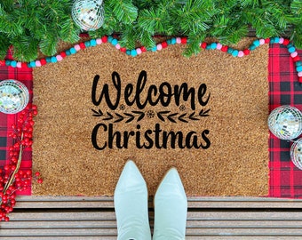 May include: A brown coir doormat with the words "Welcome Christmas" in black script. The mat is surrounded by a red and black plaid fabric, green garland, disco balls, and red berries. White boots are at the bottom.