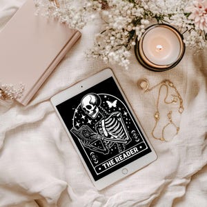 Könnte beinhalten: Ein weißes Tablet zeigt eine schwarz-weiße Illustration eines Skeletts, das ein Buch liest, mit dem Text "THE READER". Eine brennende Kerze, ein geschlossenes Buch und Goldschmuck sind auf einem cremefarbenen Stoff angeordnet, mit Blumen im Hintergrund.