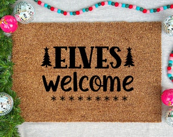 May include: A rectangular doormat with the words "ELVES welcome" in black, with Christmas tree and snowflake designs. The mat is surrounded by festive decorations, including disco balls, evergreen branches, and a colourful beaded garland.