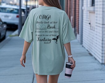 Può includere: Una maglietta oversize verde chiaro con la scritta "OMG! Becky look at her Book... It's just so BIG and Thick It must be one of those... Fantasy books" e una grafica a pipistrello. La persona tiene in mano un bicchiere rosa e nero.