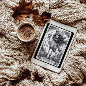 May include: A digital tablet displaying a black and white illustration of a skeleton holding a cup of coffee. The text on the tablet reads "Fall Saturdays & Pumpkin Spice Lattes". The tablet is lying on a beige knitted blanket with brown leaves.