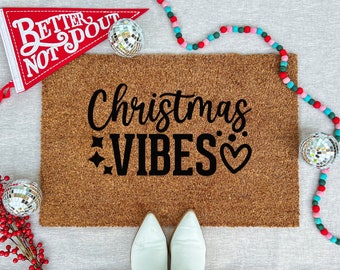 May include: A brown coir doormat with the words "Christmas VIBES" in black script, stars, and a heart. Decorative elements include disco balls, a red pennant with text, and a colourful garland. White boots are at the bottom.