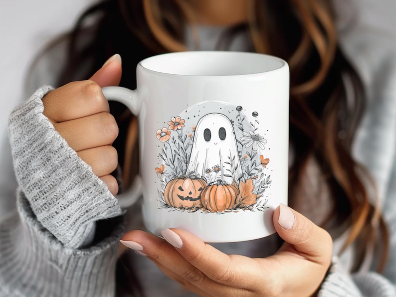 May include: White ceramic mug featuring a Halloween-themed illustration. The design includes a friendly ghost, two pumpkins, flowers, and foliage in shades of orange, gray, and white. The mug is held by a person.