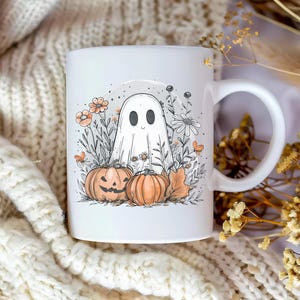 May include: White ceramic mug featuring a Halloween-themed illustration. The design includes a friendly ghost, two pumpkins, flowers, and foliage. The mug has a curved handle and is set against a cozy, textured background.