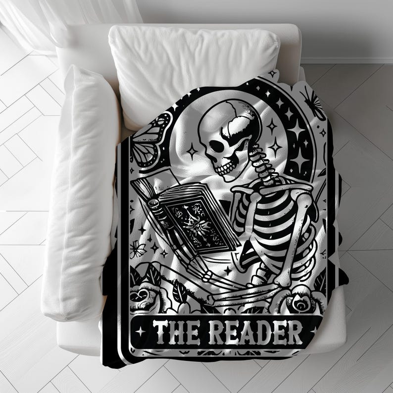Puede incluir: Manta en blanco y negro con un esqueleto leyendo un libro bajo la luna llena. El dise&ntilde;o incluye rosas, mariposas y el texto "THE READER". La manta est&aacute; sobre una cama blanca.