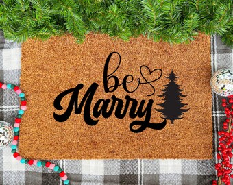 May include: A rectangular coir doormat with the words "be Merry" in black script, a heart, and a Christmas tree silhouette. The mat is surrounded by Christmas decorations, including greenery, baubles, and berries.