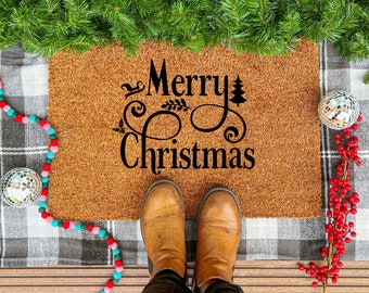 May include: A brown coir doormat with the words "Merry Christmas" in black script. The mat is surrounded by Christmas decorations, including a plaid blanket, garland, and boots.
