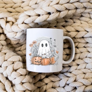 May include: White ceramic mug featuring a Halloween-themed illustration. The design includes a friendly ghost, pumpkins, flowers, and foliage. The mug has a curved handle and is set against a textured, cream-colored background.