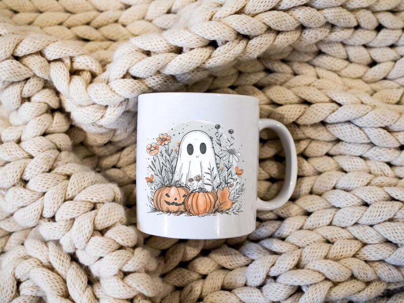 May include: White ceramic mug featuring a Halloween-themed illustration. The design includes a friendly ghost, pumpkins, flowers, and foliage. The mug has a curved handle and is set against a textured, cream-colored background.