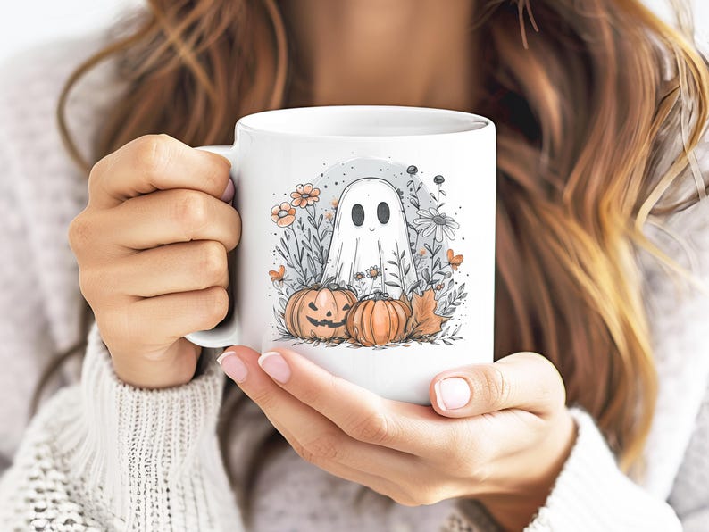 May include: White ceramic mug featuring a whimsical Halloween design. The illustration includes a friendly ghost, pumpkins, flowers, and fall leaves. The mug is held by a person.