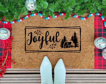 May include: A rectangular doormat with the word "Joyful" in a black script font, framed by a black rectangle. The design includes a house and trees. The mat is on a wooden surface, with red plaid fabric and Christmas decorations.