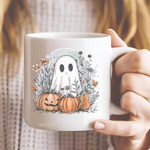 May include: White ceramic mug featuring a whimsical Halloween design. The illustration includes a friendly ghost, pumpkins, flowers, and leaves in soft orange, gray, and white tones. The mug is held by a person.