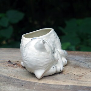 May include: White ceramic planter shaped like a sleeping cat. The planter has a small opening on the top for planting.