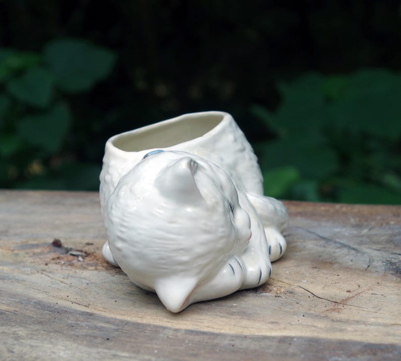 May include: White ceramic planter shaped like a sleeping cat. The planter has a small opening on the top for planting.