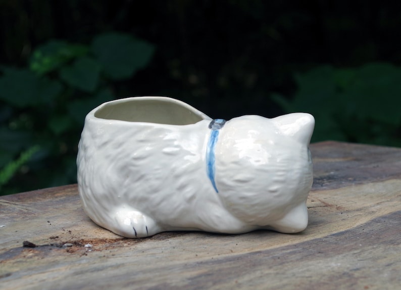 May include: White ceramic planter shaped like a cat lying down. The planter has a blue ribbon around its neck and a small opening on the top for planting.