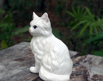 Puede incluir: Figura de gato de cerámica blanca con textura de pelaje detallada, sentada. El gato tiene ojos negros y detalles rosas en las orejas y la nariz. La figura está colocada sobre una superficie de madera desgastada, con un fondo verde borroso.
