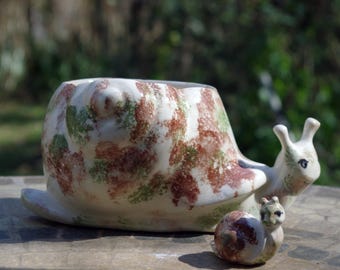 May include: A ceramic snail-shaped planter with a matching small snail figurine. The planter's shell is white with brown and green mottled patterns. The snail's head and body are off-white with small black eyes.