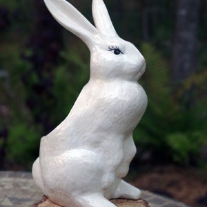 May include: A white ceramic rabbit figurine with black eyelashes. The rabbit is sitting upright with its ears perked up. It is sitting on a tree stump.