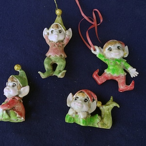 May include: Four ceramic elf ornaments with red and green clothing. The elves have pointy hats and are wearing different expressions.