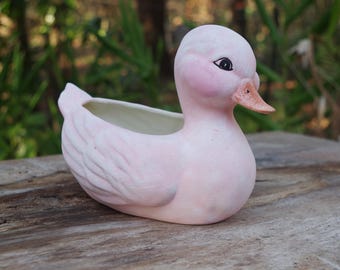 May include: A pink ceramic duck planter with a white interior. The duck has a small beak and black eyes.