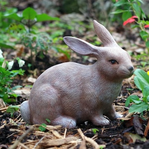 May include: A detailed, light brown rabbit statue with textured fur, seated amidst mulch and greenery. The bunny has long ears and is positioned in a natural outdoor setting, suggesting a garden or yard decoration. The statue's realistic features and colour palette create a lifelike appearance.