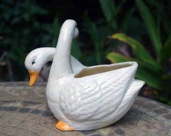 May include: White ceramic swan planter with a yellow beak and feet. The swan is in a curled position with its head tucked under its wing. The planter has a small opening in the top for planting.