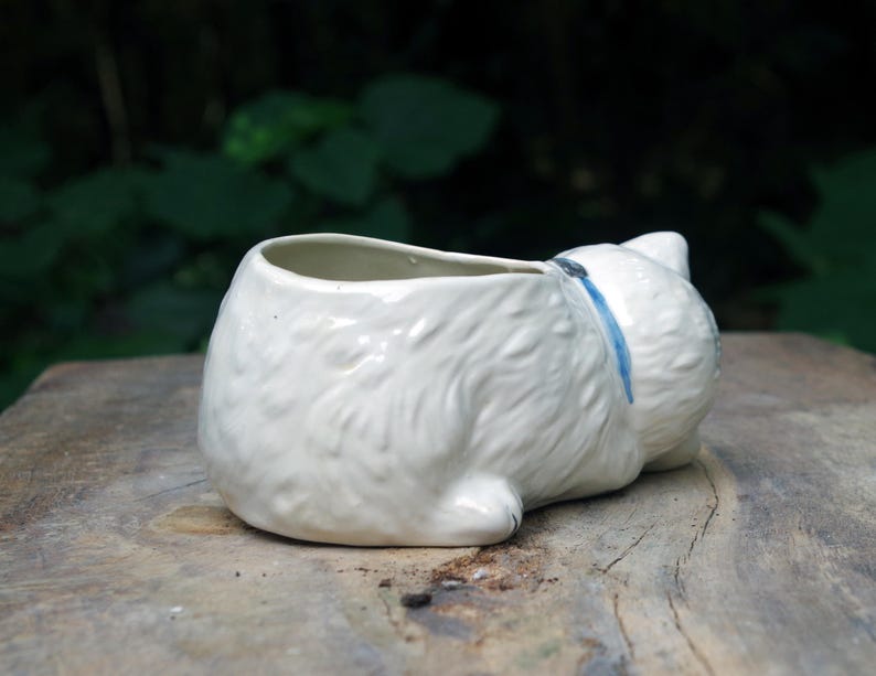 May include: White ceramic planter shaped like a cat lying down. The planter has a blue ribbon around its neck and a small opening for planting.