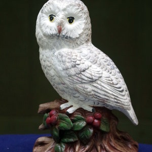 May include: White ceramic owl figurine perched on a brown tree stump with green leaves and red berries. The owl has black eyes and a yellow beak.