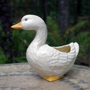 May include: A white ceramic duck planter with a yellow beak and feet. The planter features a detailed feather design and a hollowed-out area for plants. The duck is on a mosaic table with a blurred green background.
