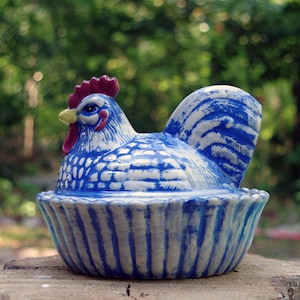 Puede incluir: Un recipiente de cerámica con forma de gallina, predominantemente azul y blanco, con cresta roja y pico amarillo. La gallina se asienta sobre una base a rayas azul y blanca a juego, creando una pieza decorativa. El fondo es verde borroso.