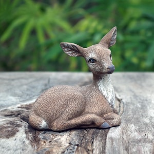 May include: A detailed figurine of a deer in a resting pose. The deer is primarily brown with a white chest and a dark nose. The figurine is set on a wooden surface with a blurred green background.
