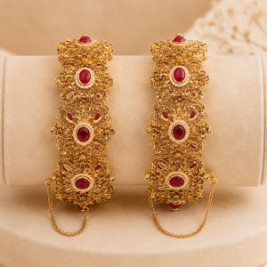 May include: A pair of gold-toned bangles featuring intricate filigree metalwork and floral-shaped motifs. Each bangle is set with multiple oval-shaped red stones surrounded by small white crystals, and includes a delicate gold chain attachment.